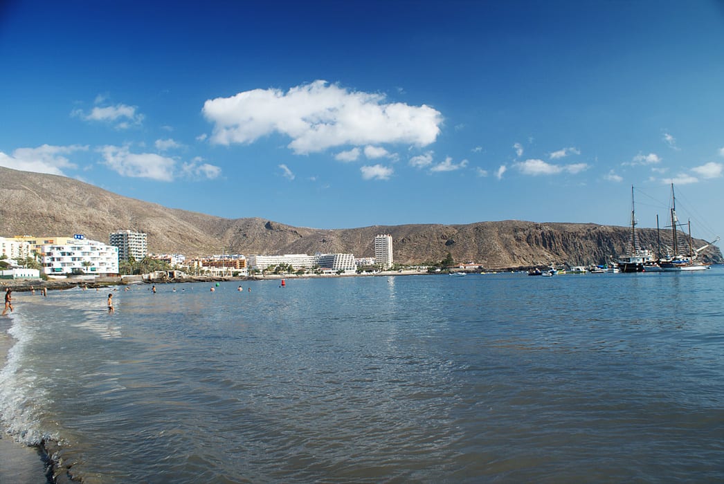 Vista panorámica de la bahía de Los Cristianos: la playa urbana del antiguo pueblo pesquero, el casco con sus edificios, los barcos junto al puerto y la montaña de Guaza.
