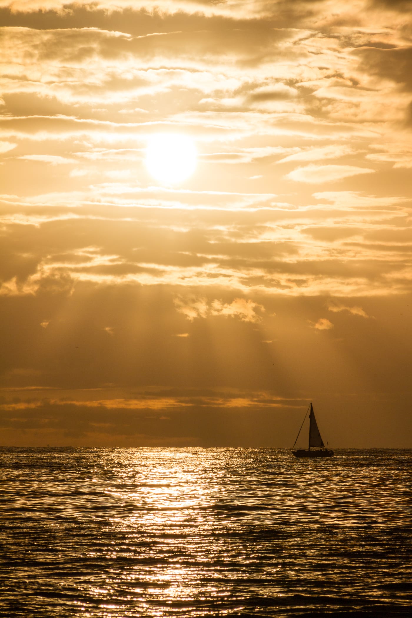 Velero navegando frente a la costa de Tenerife mientras el sol se pone sobre el mar, con sus rayos atravesando las nubes doradas.