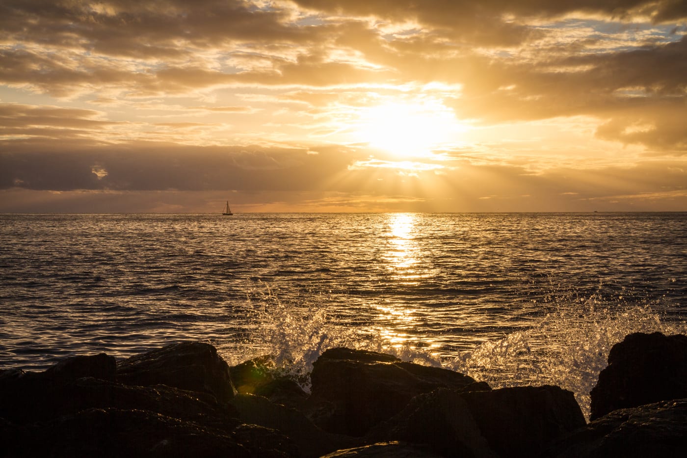 Velero al atardecer en la costa de Tenerife, con las olas rompiendo sobre las rocas en primer plano y el sol hundiéndose en el mar.