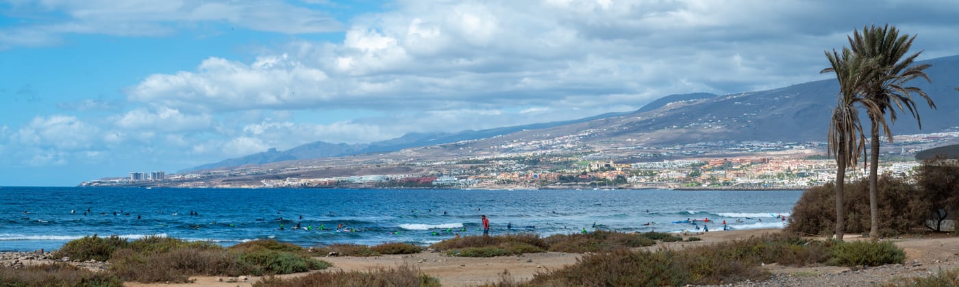 Surfistas y alumnos de surf en Playa de Las Américas, con vista hacia Playa del Duque y La Caleta, sur de Tenerife.